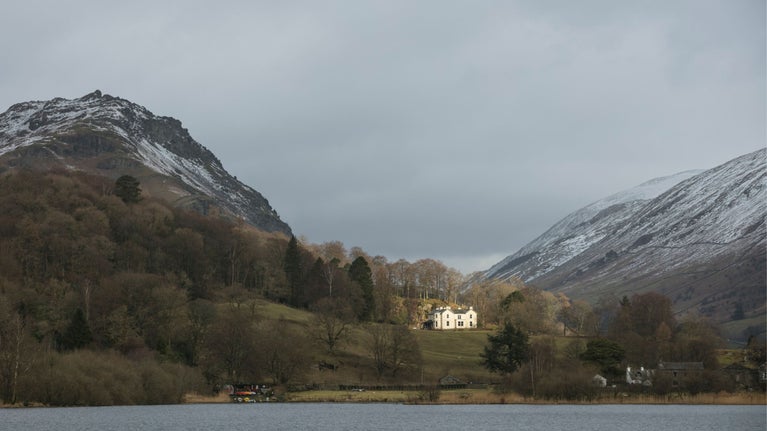 Winter scene of a house perched on hill, surrounded by higher fells and a lake in the foreground